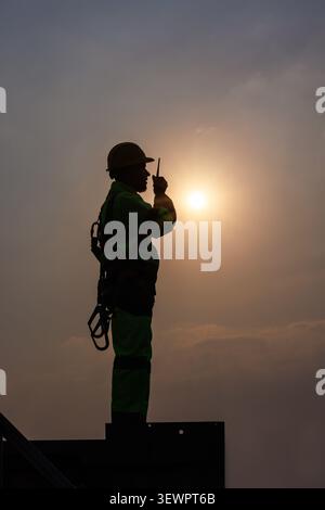 A construction worker in a hard hat and safety harness stands in silhouette using a handheld radio during a vibrant golden sunset. Stock Photo
