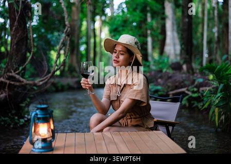 A woman relaxes at a camping table with a glass of red wine near a small creek under a lush forest canopy for a serene lifestyle concept. Stock Photo