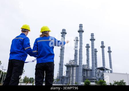 Two professional engineers wearing hard hats review blueprints while analyzing technical operations at a natural gas power plant facility. Stock Photo