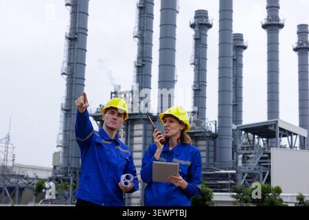 Two industrial engineers wearing safety helmets review site schematics before large refinery stacks focusing on infrastructure and energy maintenance. Stock Photo