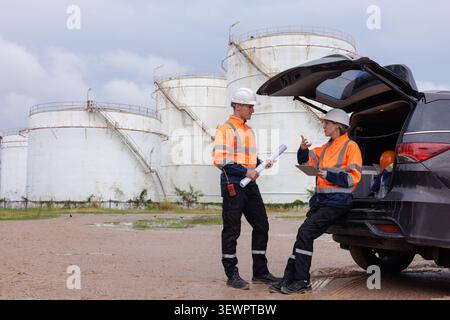 Two professional engineers in safety gear and hard hats analyze blueprints and digital data at an industrial oil storage facility. Stock Photo