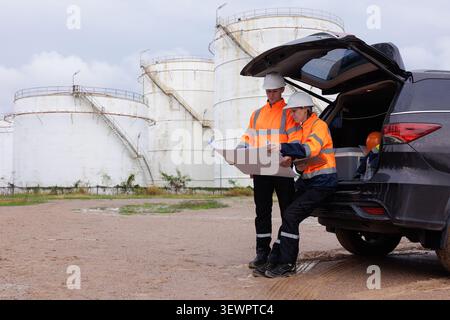 Two professional engineers in highvisibility safety gear review site plans next to an suv at an industrial bulk storage facility. Stock Photo