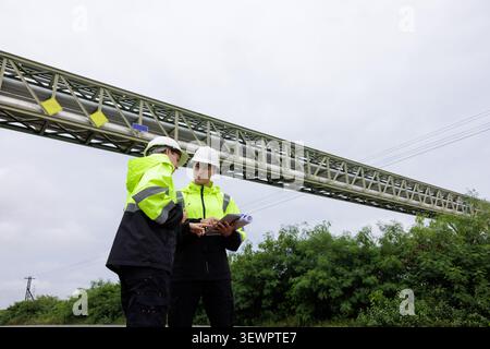 Two engineers in safety gear collaborate on a construction site inspection reviewing project blueprints on a digital tablet near industrial infrastruc Stock Photo
