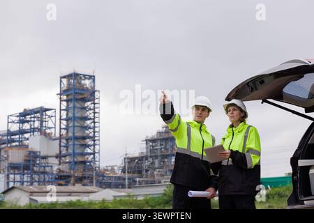 Two industrial engineers wearing hard hats and reflective gear discussing construction project details on a tablet at a chemical plant. Stock Photo
