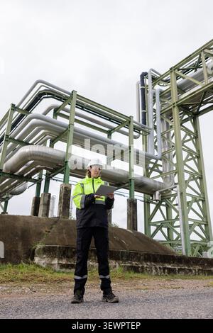 A professional female engineer in safety gear uses a tablet to inspect infrastructure at an industrial chemical processing facility. Stock Photo