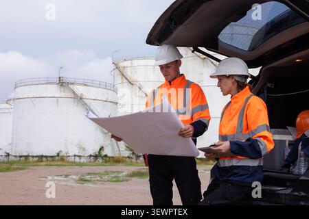A male and female engineer wearing safety gear examine blueprints beside a vehicle at a large fuel storage depot to coordinate site maintenance. Stock Photo