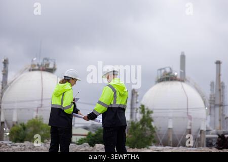 Two professional engineers in high visibility gear and hardhats review construction blueprints at a large petrochemical refinery plant. Stock Photo