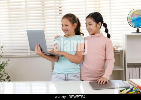 Two elementary age girls stand together smiling while interacting with a laptop during a study session in a bright home office. Stock Photo