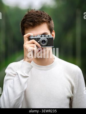 Young man in white sweater holding vintage camera in front of his face. Male photographer taking picture outdoors Stock Photo