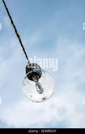 A close-up, low-angle shot of a clear, round light bulb covered in raindrops, hanging by a wire against a bright, cloudy sky. Stock Photo