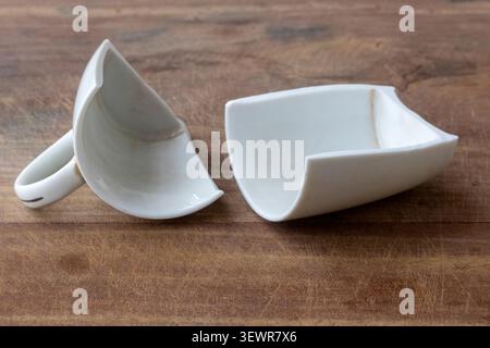 Close-up of a broken white ceramic tea cup in two pieces on a rustic wooden table surface. Ideal for abstract concepts of failure, broken relationship Stock Photo