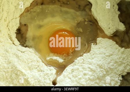 Raw egg yolk and white sitting in a mound of light brown flour, preparing ingredients for baking or pasta making in a close up view, highlighting the Stock Photo