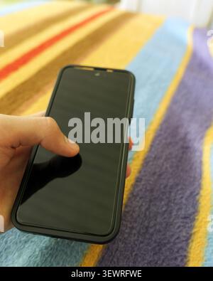 Hand holding a black smartphone with a blank screen, pointing a finger on the display, interacting with mobile digital technology over a colorful stri Stock Photo