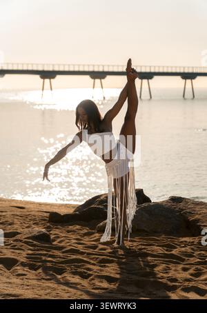 Beautiful Cuban dancer performing a standing split pose on the sand at sunrise on El Coco beach in Badalona with the Pont del Petroli in the backgroun Stock Photo