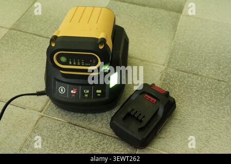 Yellow and black lithium ion battery charging in a black power tool charger with indicator lights. Another battery pack resting on the tiled floor bes Stock Photo