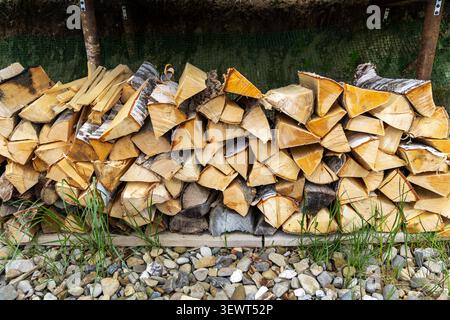 Stacked Firewood Under Outdoor Shelter. Stacked split firewood under an outdoor shelter with birch bark, rough log faces, green grass, and gravel Stock Photo