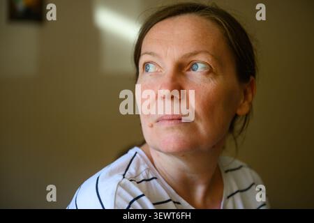 Mature woman with a neutral expression standing against a plain wall in soft light. Authentic portrait representing everyday life, resilience and intr Stock Photo