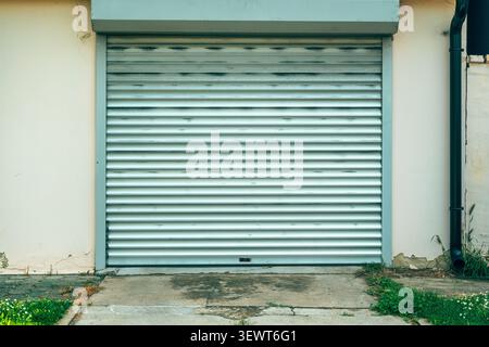 Closed metal garage door with horizontal rolling shutter panels sits in a weathered suburban wall above a cracked concrete driveway, framed by a drain Stock Photo