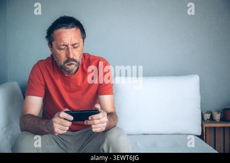 Serious mature man using smartphone at home, browsing online content, messaging or consuming digital media, selective focus Stock Photo
