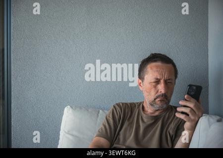 Middle-aged man looking thoughtfully at smartphone while sitting on sofa, selective focus. Stock Photo