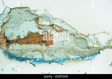 Peeling plaster reveals exposed brick, crumbling mortar and a torn blue mesh on a weathered wall, offering gritty texture and urban decay for backgrou Stock Photo