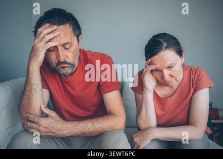 A middle-aged couple sit side by side on a sofa, both looking worried and withdrawn as they face stress, emotional distance and relationship strain in Stock Photo