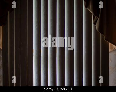 Slices of bright daylight peeking through closed vertical window blinds inside a dark room with a fabric curtain valance Stock Photo