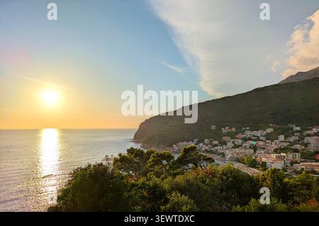 A high-angle view shows the bright sun setting over the sea, lighting up the coastline, green hills, and a dense coastal town in Petrovac, Montenegro. Stock Photo