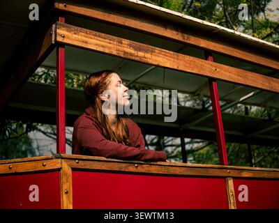 Young woman with red hair sits on a vintage trolley. She wears a maroon top, looking up with a content smile. The red trolley features wooden panels a Stock Photo