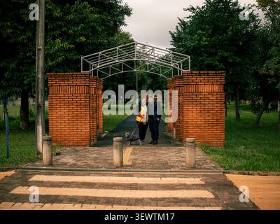 Red brick archway frames a metal gateway structure. Lush green trees fill the park background. Two people stand close under the arch. They smile, sugg Stock Photo