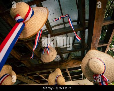 Straw hats hang from a wooden pergola. Red white blue ribbons adorn each hat. Mesh roof covers rustic wooden beams. Festive decorations suit celebrati Stock Photo