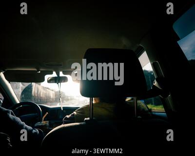 Dark car interior. Driver's hands rest on the steering wheel. Sunlight streams through the windshield. Blurred road shows motion ahead. Back seat feat Stock Photo