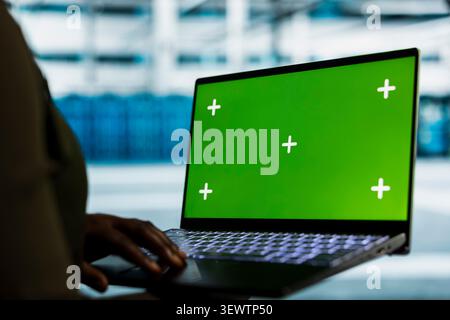 Software developer uses a green screen laptop in a data center for hardware diagnostics, screen replacement, and server workflow monitoring. Stock Photo