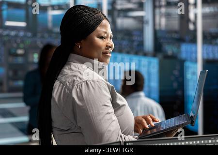 Happy technician uses a laptop in a server room to manage data backups, resolve technical issues and support reliable recovery. Stock Photo