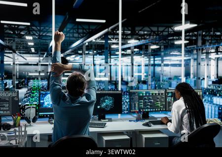 Software developer stretches at a workstation in a server hub to relieve back fatigue after long technical work. Stock Photo