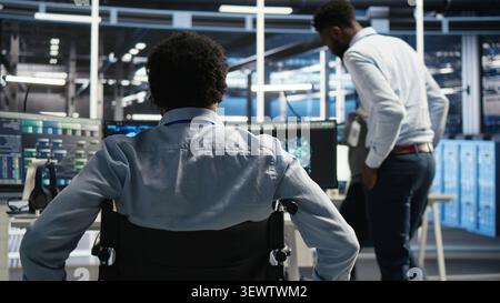 Data infrastructure center software programmer in wheelchair arriving at the office operates AI visual programming workflow to minimize system downtime. Stock Photo