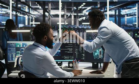 Computing hub team supervisor checking AI generated data analytics graphs, congratulating staff member, while team leader sharing a fist bump with system admin working with AI showing system metrics. Stock Photo
