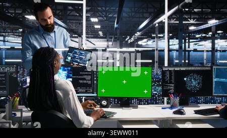 Female software programmer talking with team supervisor in high tech server room, checks AI diagnostics on chroma key computer. Stock Photo
