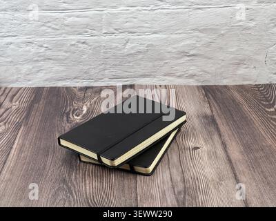 Two professional black journals with elastic closures stacked on a rustic wooden surface against a white brick wall. Stock Photo