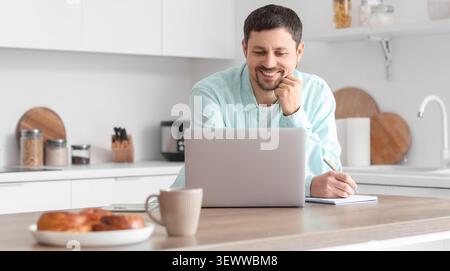 Man working on laptop and taking notes in kitchen Stock Photo