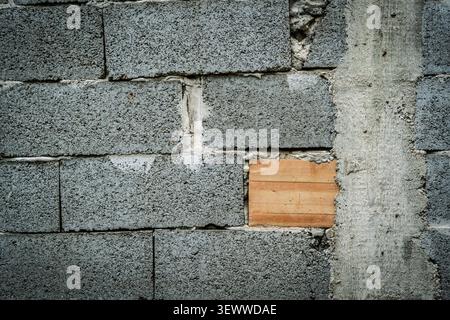 Close view of a rough wall made from grey concrete blocks, with one smooth orange brick and an unfinished vertical concrete patch creating contrast in Stock Photo