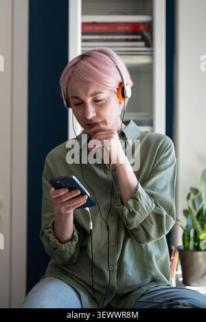 Interested thoughtful mature woman enjoying listening to music and podcasts online using mobile app. Stock Photo