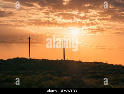 Sunset over rural landscape with power lines silhouetted against golden sky. Stock Photo