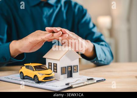 Insurance concept life home auto protection shown through male hands shielding house model and small yellow car on office desk, providing financial se Stock Photo