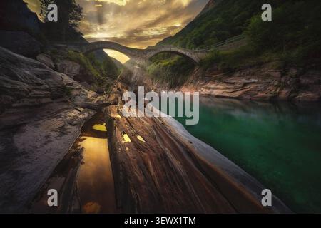 View of the historic stone arch bridge Ponte dei Salti spanning the turquoise Verzasca River with smooth rock formations and golden sunset light in Sw Stock Photo