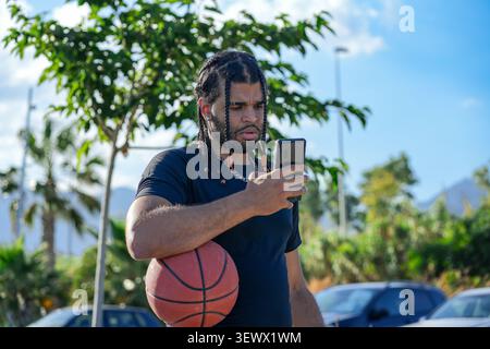 Young man checking phone while holding basketball outdoors Stock Photo