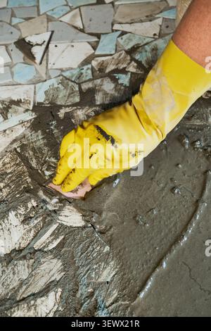 Gloved hand laying pieces of reclaimed broken ceramic tiles into cement mortar. Upcycling old materials. DIY mosaic floor renovation Stock Photo