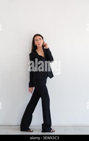 Woman looking up in black formal pantsuit or suit stands against white wall in studio. Caucasian brunette model with long hair. Business portrait Stock Photo