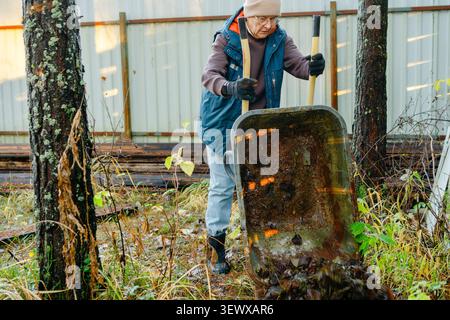 Senior woman standing with shovel behind wheelbarrow in backyard outdoors. Elderly homeowner holds garden tools ready for yard maintenance work on the property. Active retiree equipped for seasonal Stock Photo