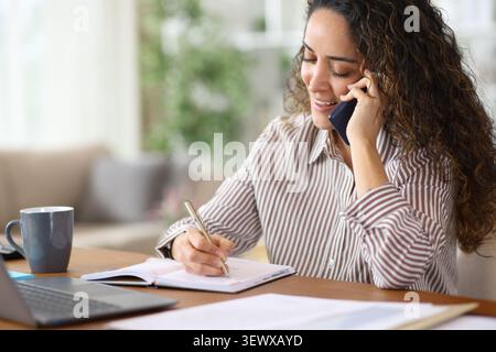 Happy adult teleworker writing in agenda and talking on phone while working at home Stock Photo